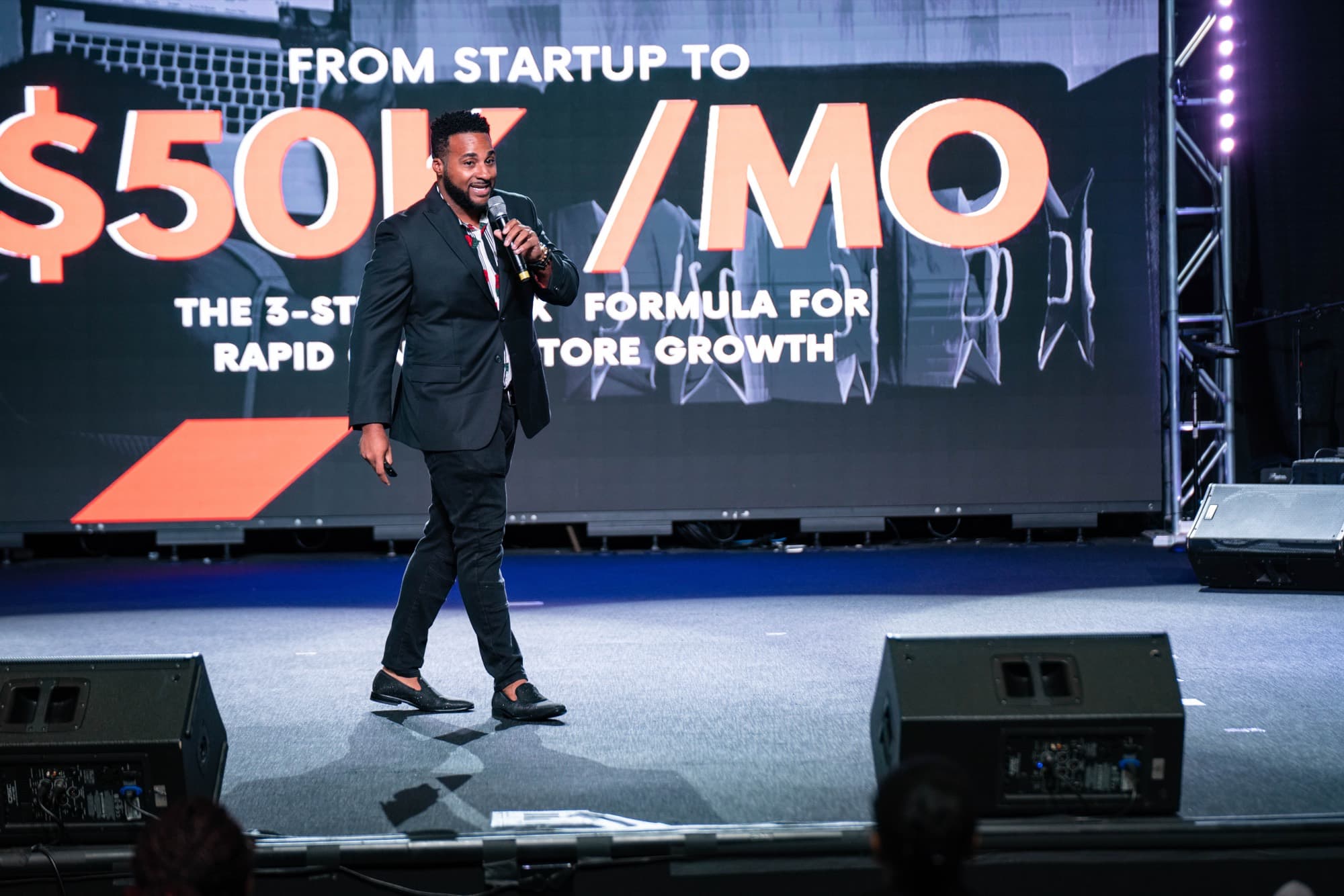Mike Eyo speaking on stage in front of a screen reading “From startup to $50K/mo — the 3-step formula for rapid store growth”